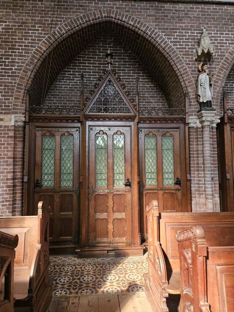 Houten biechthokjes in de Bonifatiuskerk in Leeuwarden, met glas-in-loodruitjes en gotische details, ingebouwd in een bakstenen muur. Op de voorgrond staan houten kerkbanken en op de vloer liggen rijk versierde tegels. Rechts is een heiligenbeeld te zien.