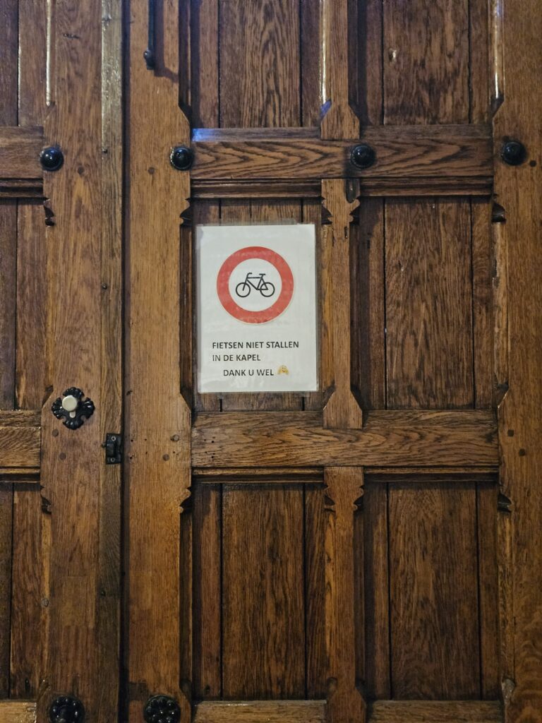 Houten kerkdeur in de Bonifatiuskerk in Leeuwarden met een A4-bordje dat fietsen verbiedt. Op het bord staat een rood verkeersbord met fiets-icoon en de tekst: “Fietsen niet stallen in de kapel, dank u wel”.