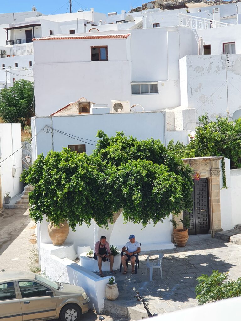 Twee mannen zitten in de schaduw onder een grote boom op een plein in Lindos.