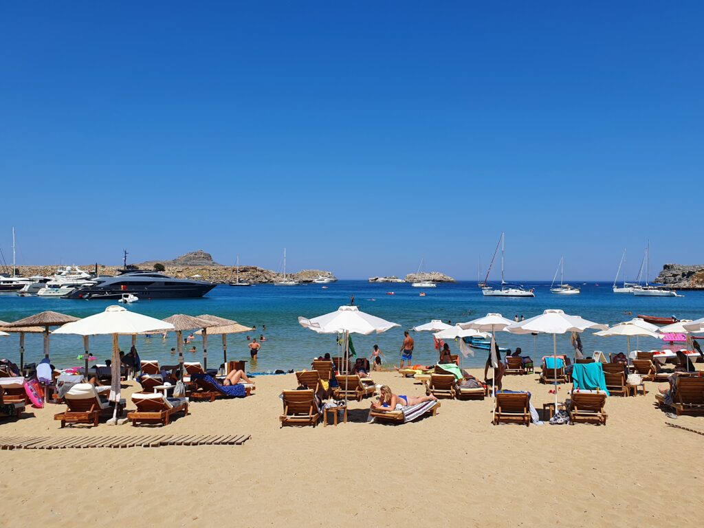 Strand bij Lindos met ligbedden, parasols en uitzicht over de helderblauwe Egeïsche Zee.
