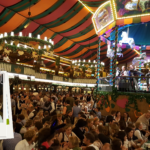 Een volle Oktoberfest-tent in München met een liveband op het podium, terwijl een selfie van een enthousiaste bezoeker in traditionele Beierse kleding als fotoframe in beeld is geplaatst.