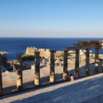 Rij stenen zuilen op de acropolis van Lindos, met bezoekers en de blauwe zee op de achtergrond.