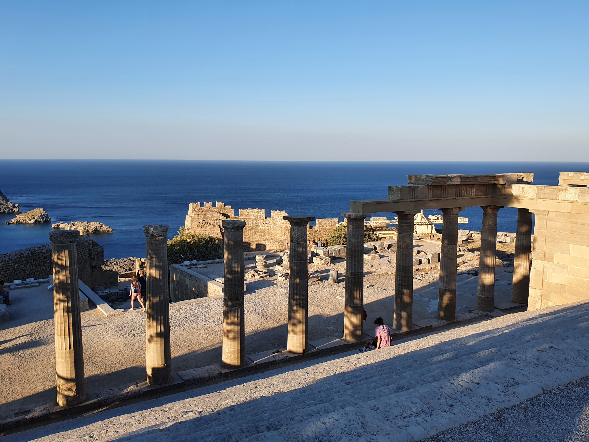 Rij stenen zuilen op de acropolis van Lindos, met bezoekers en de blauwe zee op de achtergrond.
